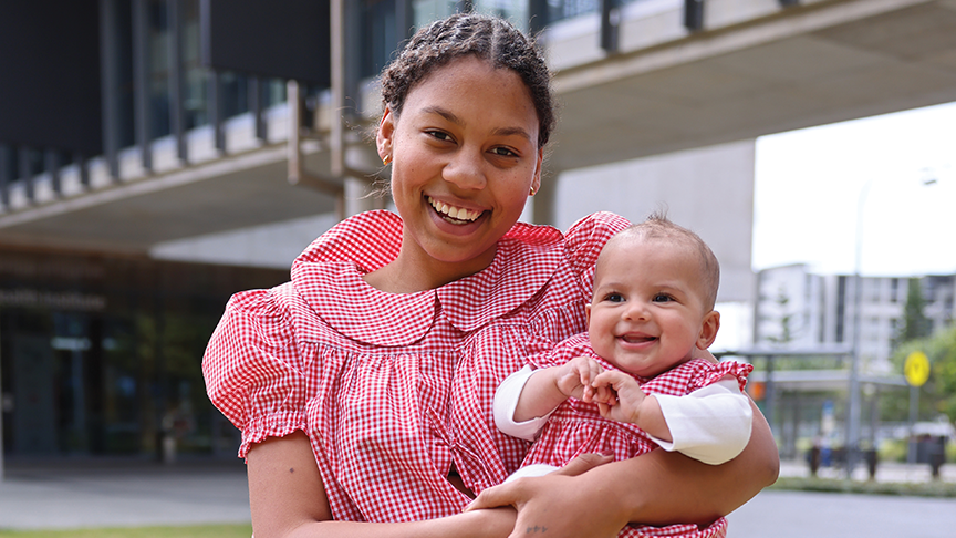 A woman stands smiling holding a young smiling toddler, wearing matching red checked tops, outside Sunshine Coast University Hospitaloutside