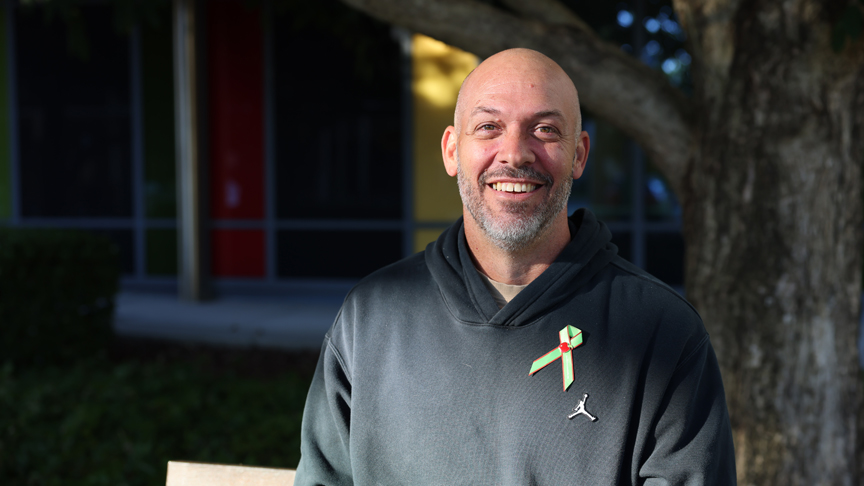 Headshot of a man, wearing a black hoodie with a green ribbon pin, sitting on a bench, smiling in front of a tree