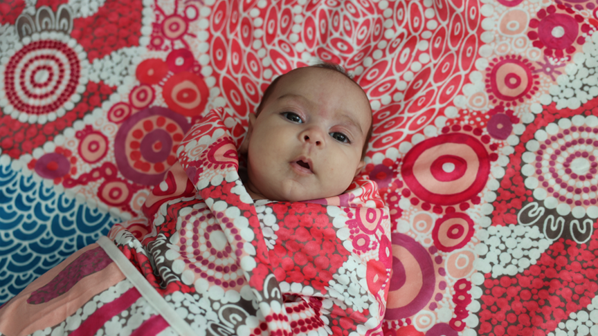 A baby wrapped in a blanket printed with Aboriginal and Torres Strait Islander artwork, lying on a blanket with the same print.