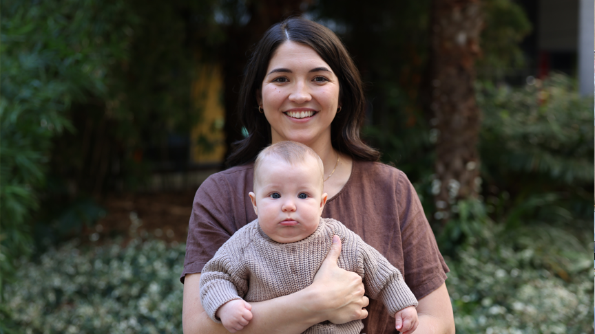A woman smiles holding a baby, standing in front of trees and greenery