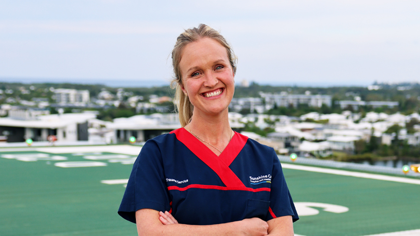 A headshot of a female nurse in dark blue scrubs smiling with arms folded, standing on a green helipad on top of a building. 