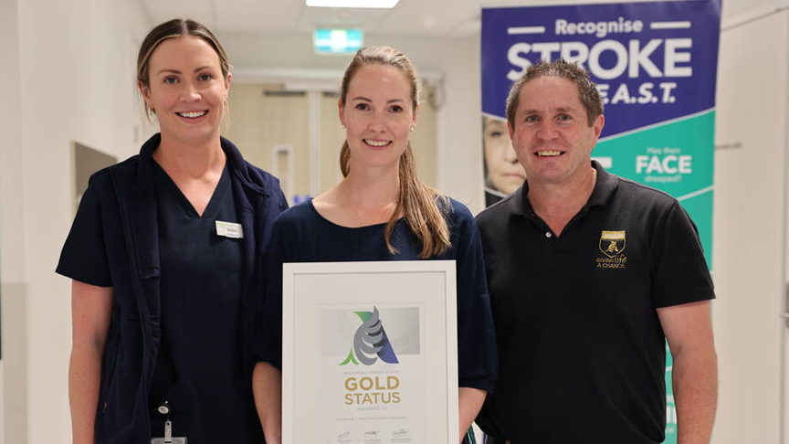 3 people standing in a hospital hallway. The person in the middle is holding a framed certificate that has the words 'Gold status' written on it.