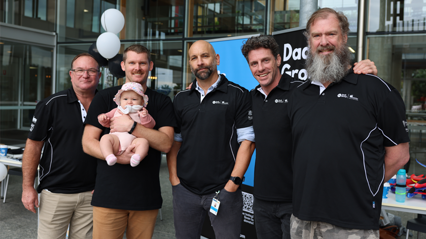 A group of 5 men standing outside Sunshine Coast University Hospital. One of the men is holding a baby in his arms.