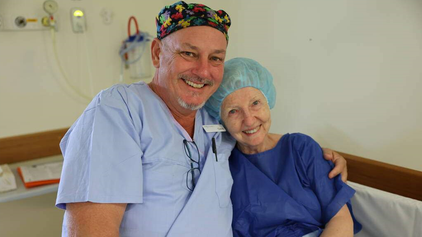 A man wearing blue scrubs and a coloured scrub hat smiles with his arm around a smiling lady in a blue hospital gown and hair net. 