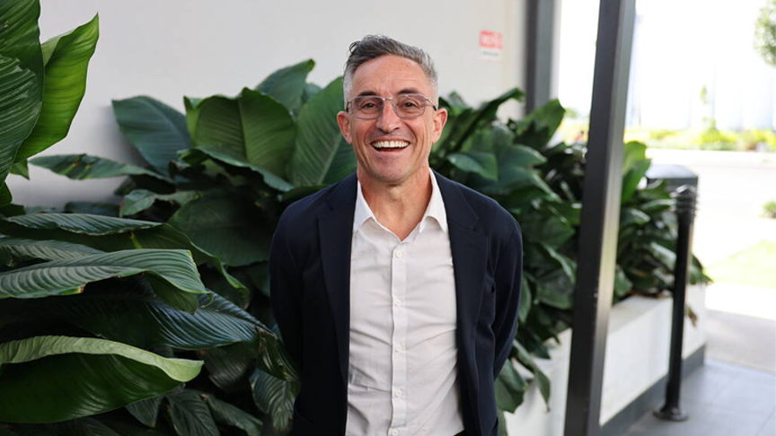 A man in a dark suit with a white button shirt wearing glasses stands smiling with his hands behind is back, in front of some green foliage against a white wall.