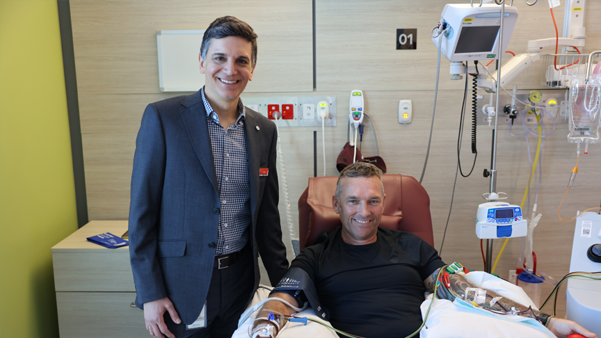 A man sat in a chair in hospital with wires and tubes connected to his arms as another man stands next to him