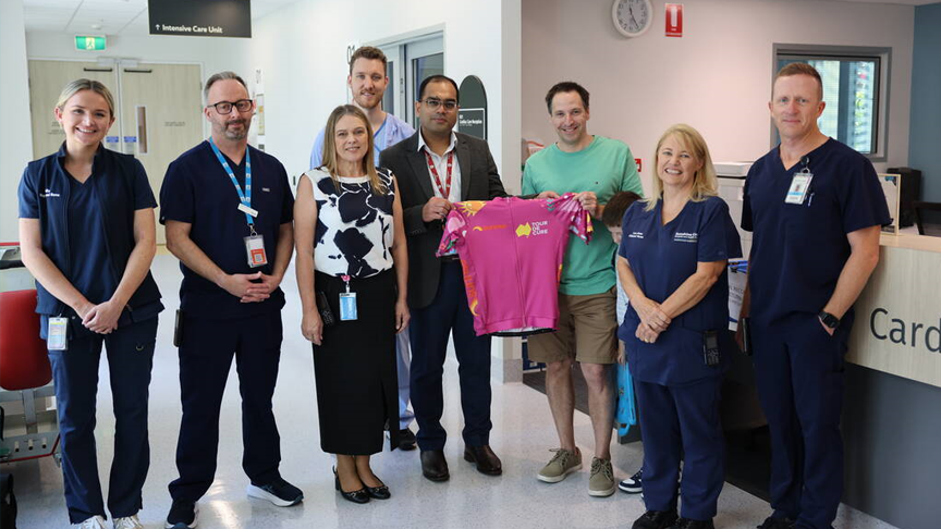 A group of people standing in a hospital reception area. Two people in the middle hold a pink cycling jersey.