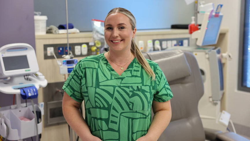 A nurse wearing green scrubs stands smiling in a renal treatment room