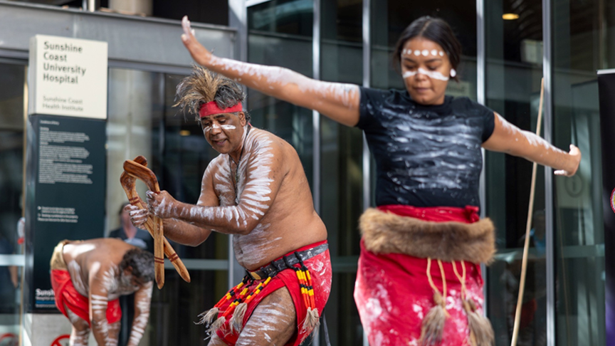 A group of Aboriginal dancers in colorful traditional attire and with ochre-painted bodies performing a traditional dance outside the front of Sunshine Coast University Hospital.