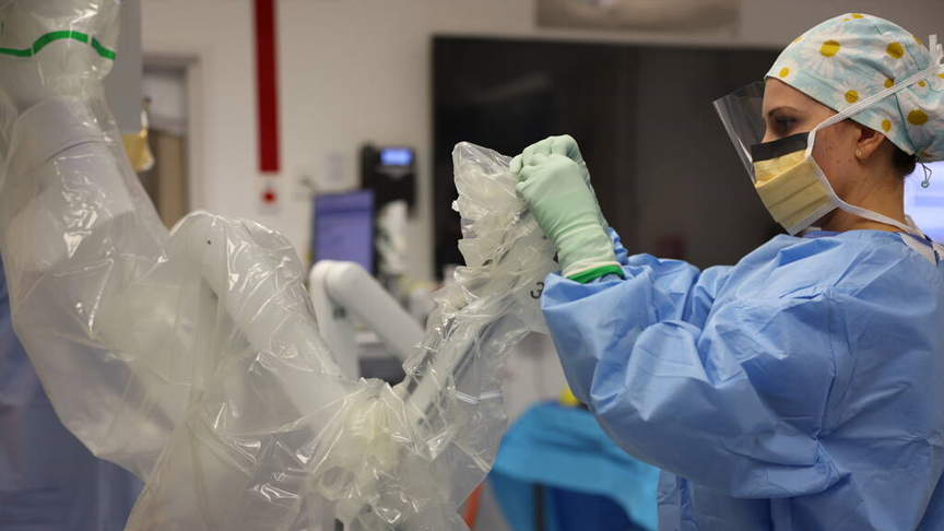 A nurse dressed in surgical gown, and full PPE working on a piece of robotic equipment covered in plastic.