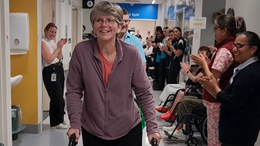 A woman using a wheely-walker to walk down a hospital corridor, lined with people smiling and clapping
