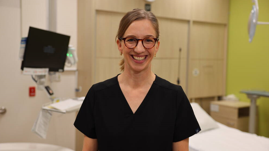 Headshot of a woman in black scrubs and wearing glasses, smiling in a hospital room with a hospital bed and equipment in the background.