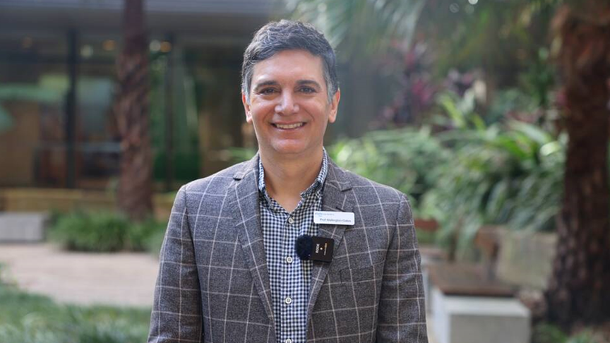 Headshot of a man smiling, while standing outside with greenary and trees in the background, with a namebadge and microphone on his jacket collar