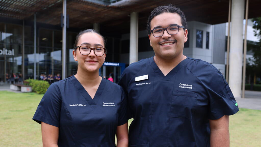Headshot of 2 people standing on the grass in the courtyard outside Sunshine Coast University Hospital smiling in dark blue nursing scrubs