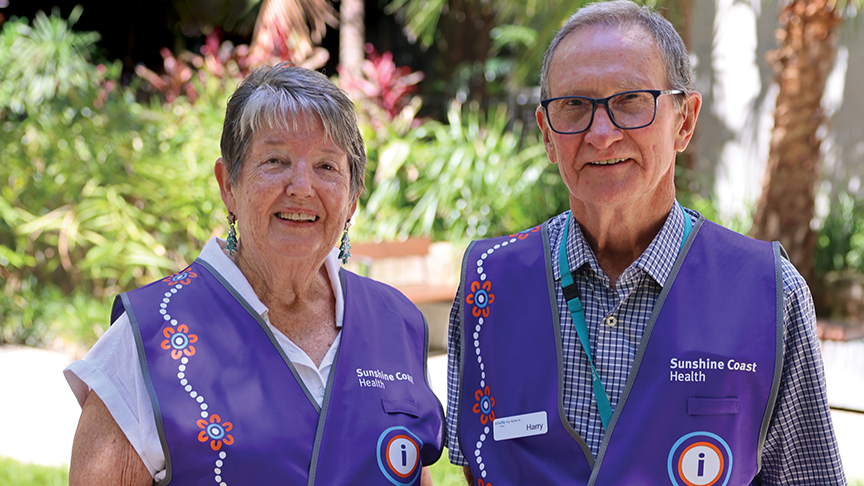 2 older persons stand outside Sunshine Coast University Hospital wearing purple vests.