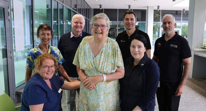 A woman in a light green dress stands smiling surrounded by a group of 6 hospital workers