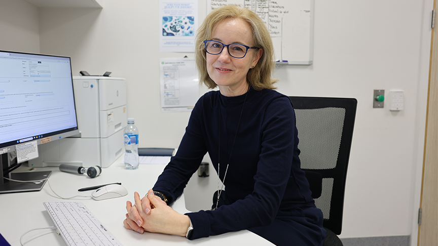 Doctor Leith Banney wearing glasses and dressed in black sits at a desk in front of a computer screen in a hospital office.