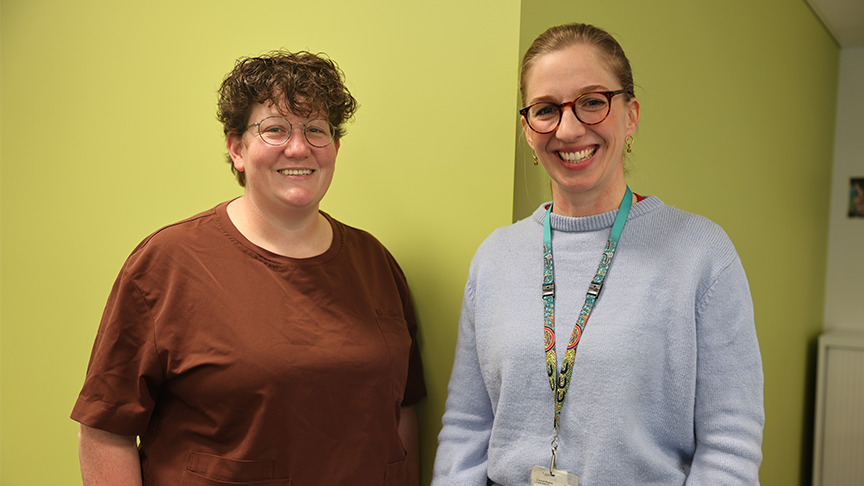 Two women, both smiling and wearing glasses stand against a green painted wall.